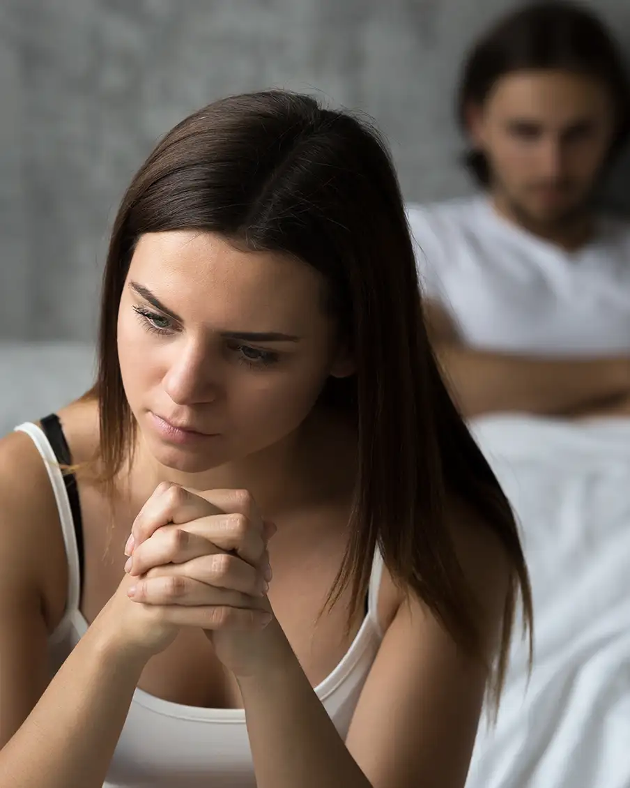 Eine Frau sitzt nachdenklich mit gefalteten Händen im Vordergrund, im Hintergrund sitzt ein Mann auf einem Bett. Beide tragen weiße Oberteile. Die Szene deutet auf eine nachdenkliche oder ernste Stimmung hin.