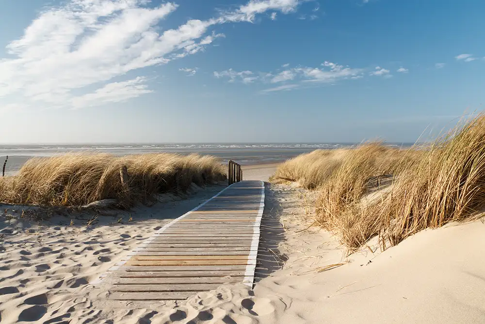 Holzsteg führt durch Sanddünen zu einem Strand. Dünengras säumt den Weg. Der Himmel ist blau mit vereinzelten Wolken. Im Hintergrund sind das Meer und der Horizont sichtbar.