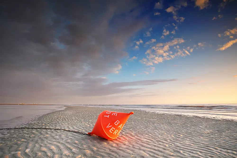Eine orangefarbene Boje liegt auf dem sandigen Strand, der von Wasser umgeben ist. Der Himmel ist teils bewölkt mit blauem und orangem Licht während der Dämmerung. Eine Kette verbindet die Boje mit dem Boden.