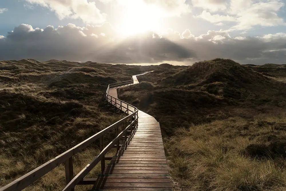 Ein hölzerner Spazierweg schlängelt sich durch eine grasbewachsene Dünenlandschaft unter einem bewölkten Himmel. Die Sonne strahlt hell durch die Wolken und wirft Licht auf den Weg und die Hügel.