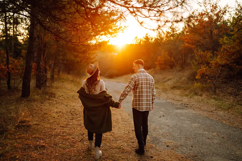 Ein Paar geht auf einem Waldweg dem Sonnenuntergang entgegen. Beide tragen Herbstkleidung. Links trägt die Person einen Hut, beide halten sich an den Händen. Bäume säumen den Weg, das Laub ist herbstlich gefärbt.